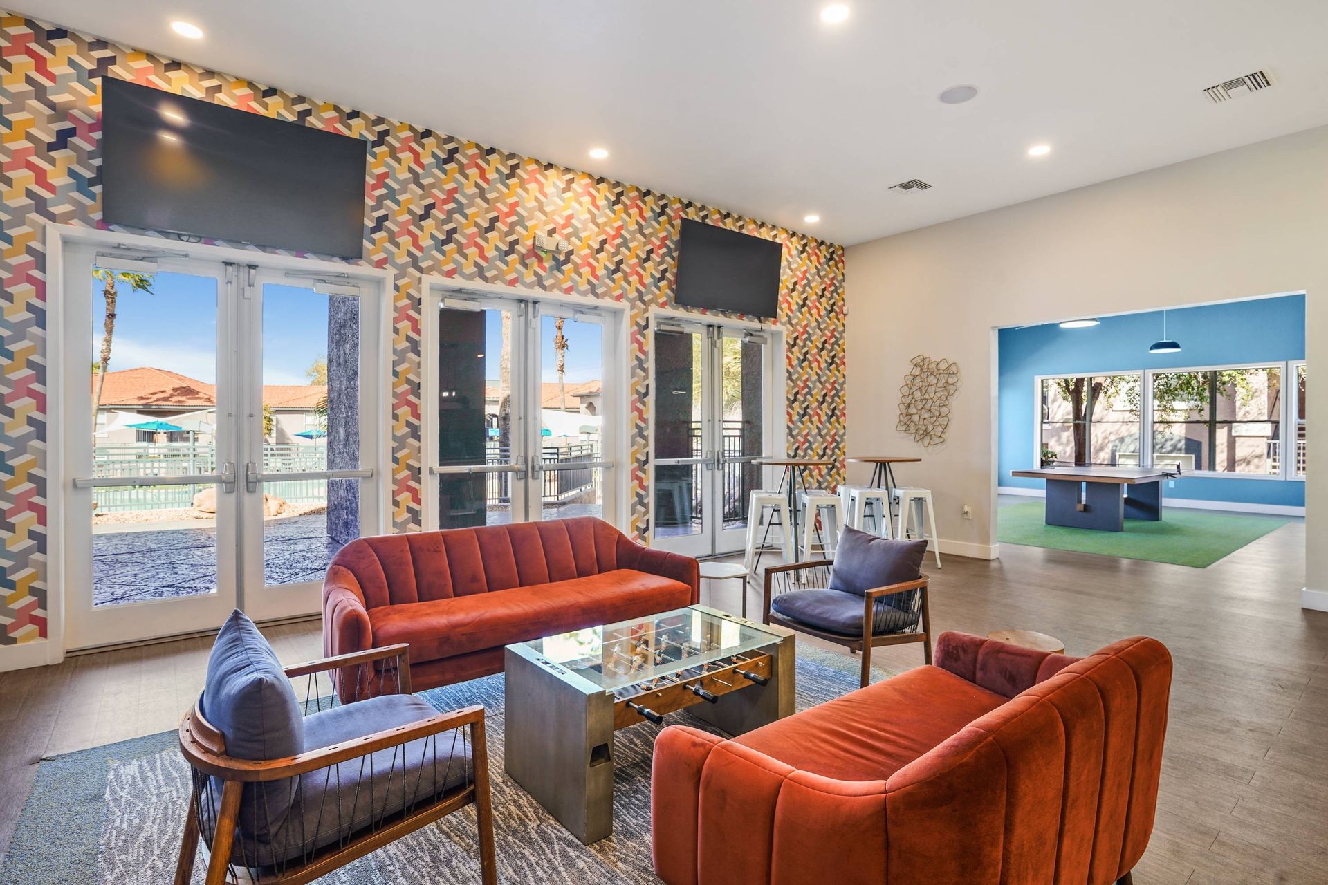 Communal lounge with colorful geometric wallpaper, glass exterior doors, wall-mounted TVs, and orange seating.