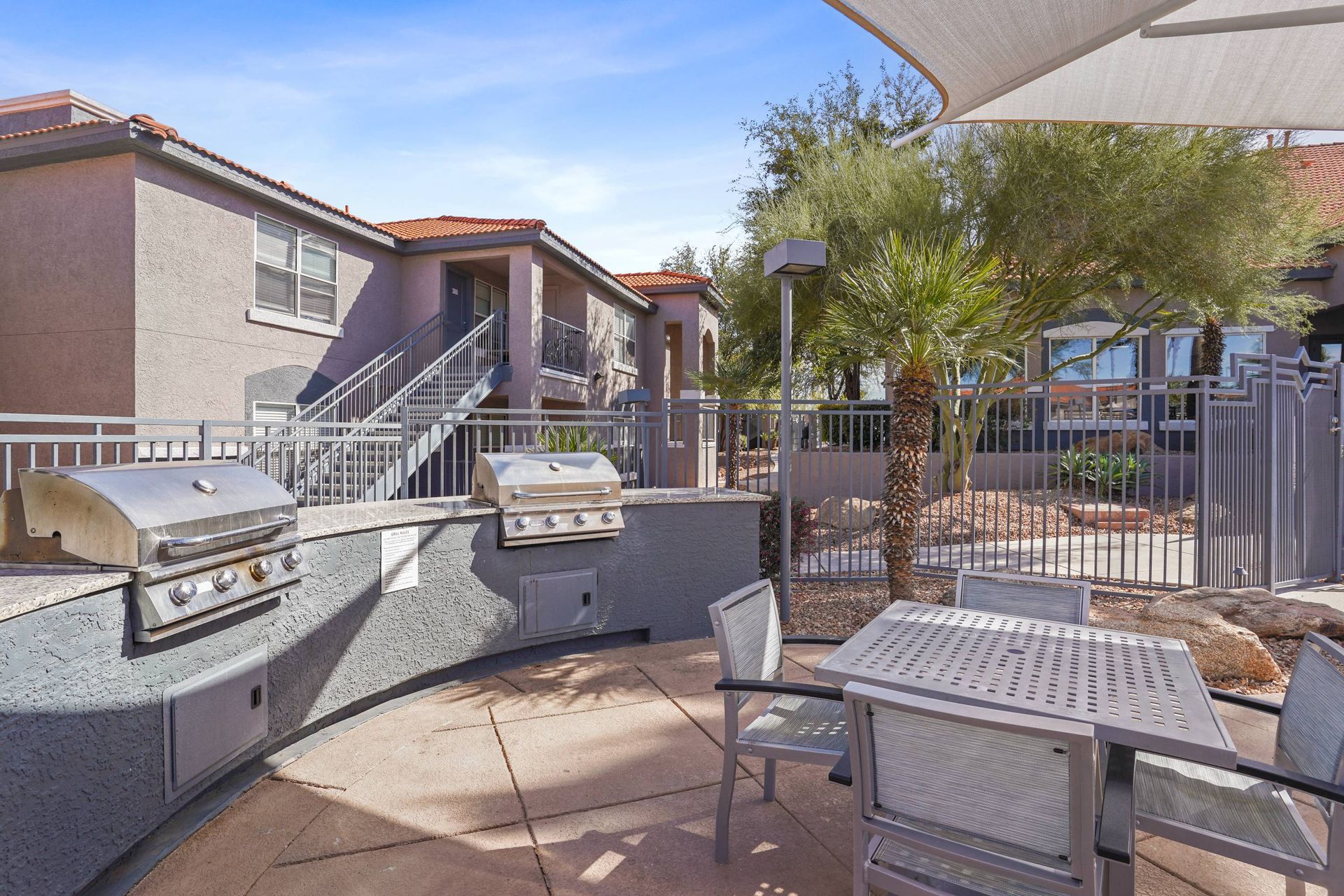 Outdoor communal grilling area with table, chairs, and palm trees in a gated apartment courtyard.