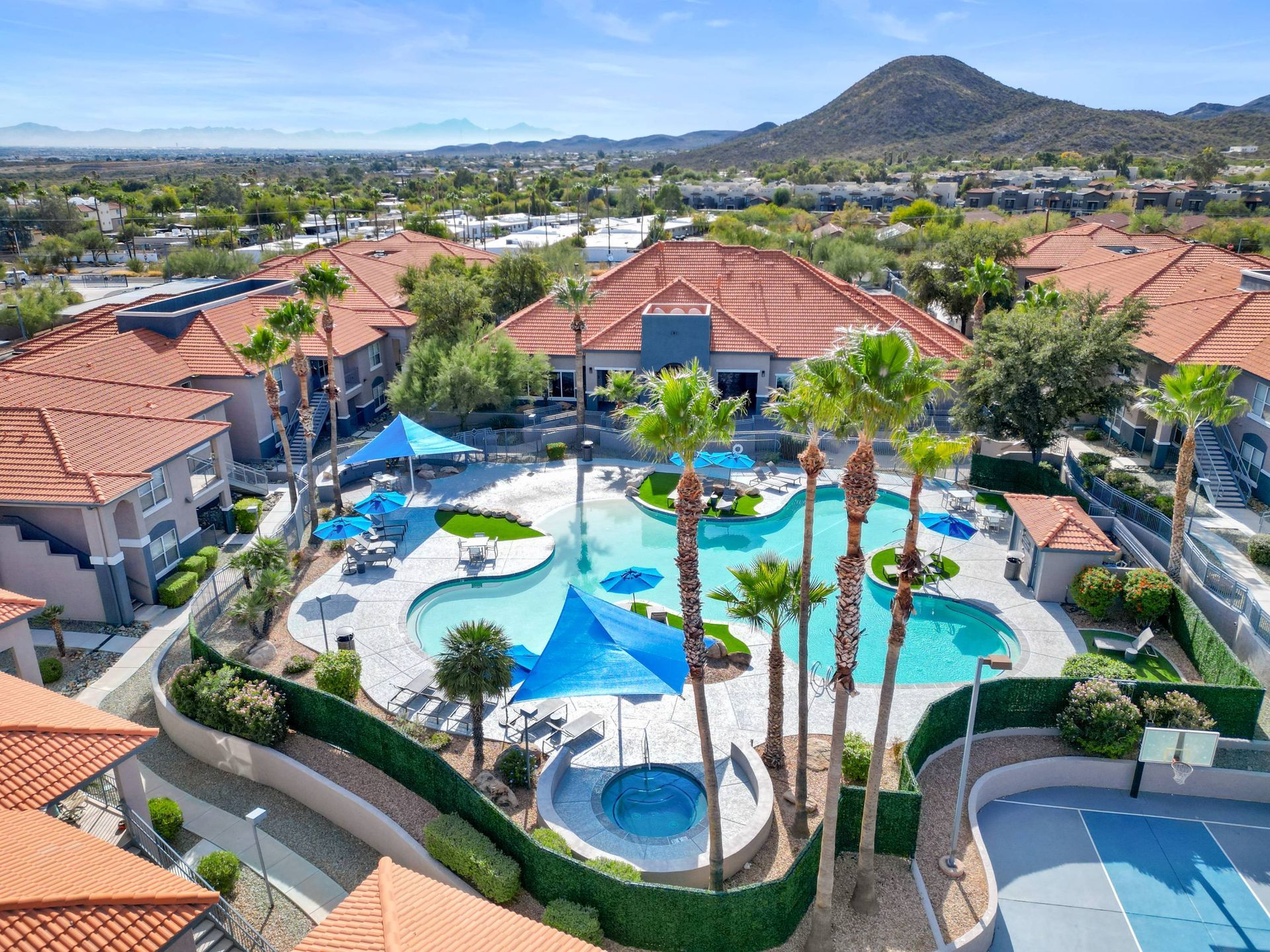 Aerial view of a resort-style apartment community pool area with palm trees and blue umbrellas.