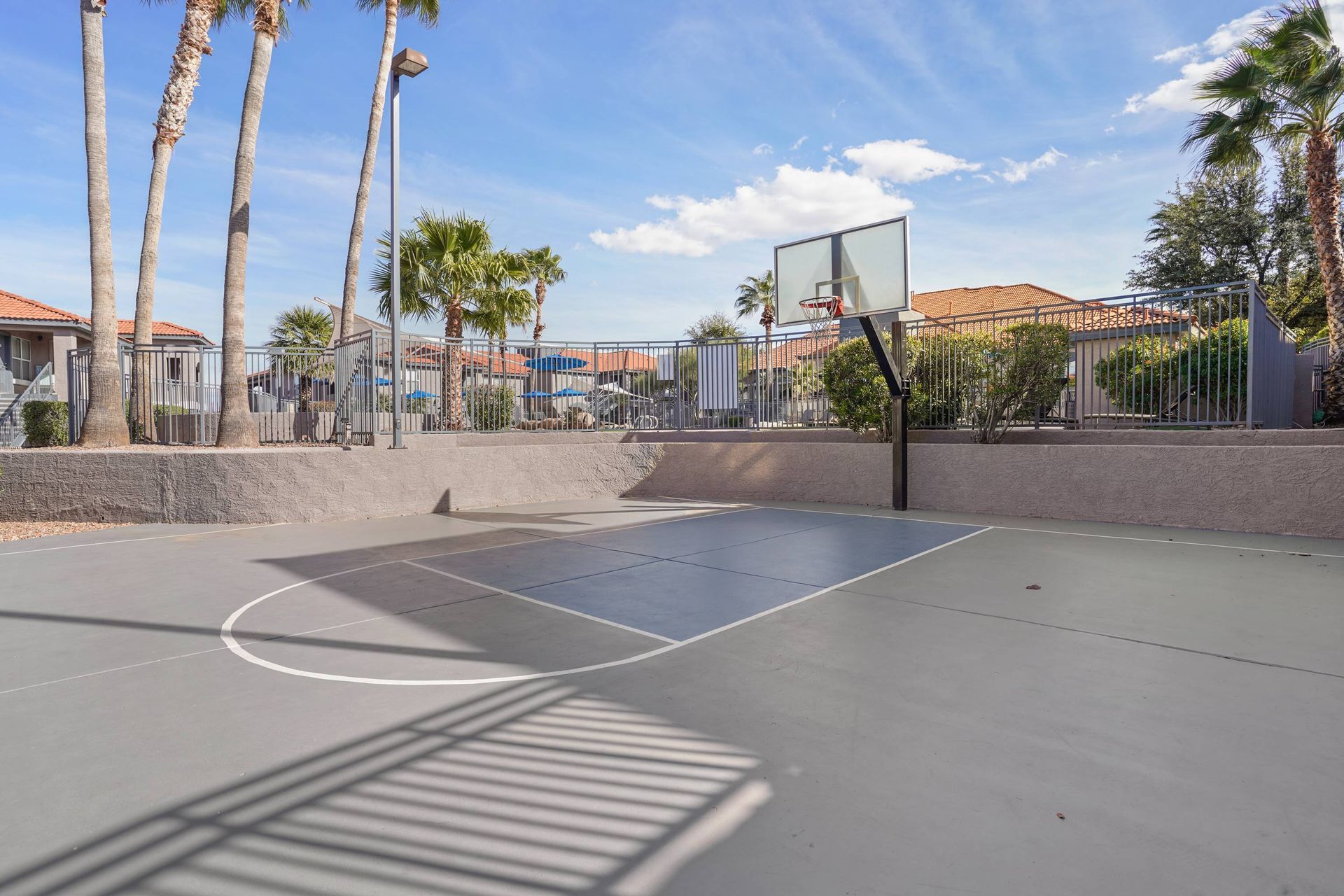 Outdoor basketball court with hoop, palm trees, and a surrounding fence.