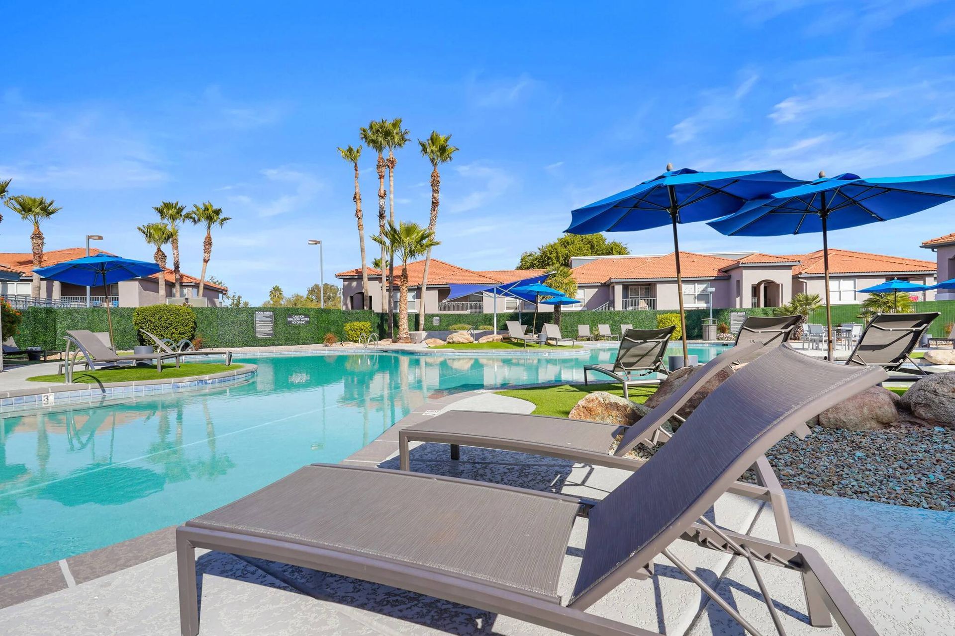 Outdoor apartment pool with lounge chairs, blue umbrellas, and palm trees under a clear blue sky.