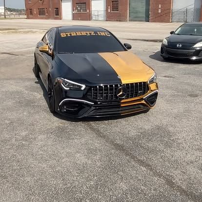 Black and gold Mercedes-Benz car parked on asphalt with a second car behind it and a brick building in the background.