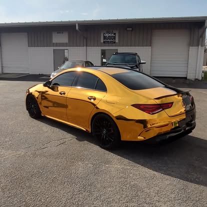 Gold-wrapped car parked in front of a building with garage doors. Black wheels and tinted windows. Sunny day.