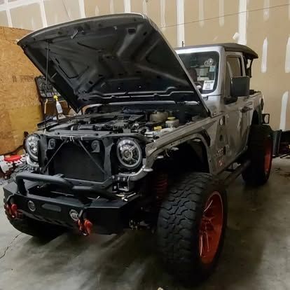 Silver Jeep Gladiator with the hood open, parked indoors. It has large tires and a black bumper.