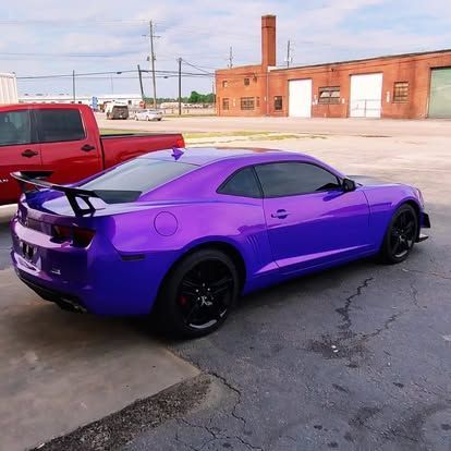 Purple sports car with black wheels and spoiler parked in front of red brick building and truck.