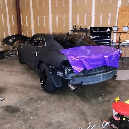 Black car in a garage with purple wrapping on the rear, being worked on.