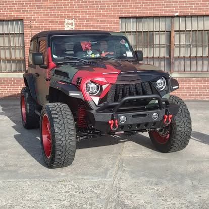 Camouflaged Jeep with red accents and large tires parked in front of a brick wall.