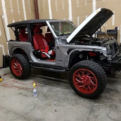 Silver Jeep with red interior and wheels, hood open, in a garage.