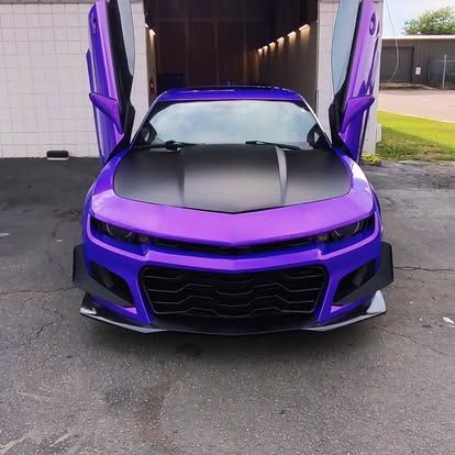 Purple sports car with doors open upward, black hood, in front of a building.