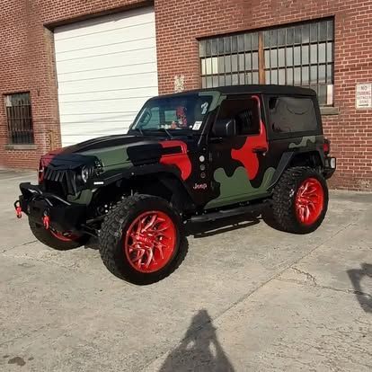 Camouflaged Jeep with red accents and wheels parked in front of a brick building.