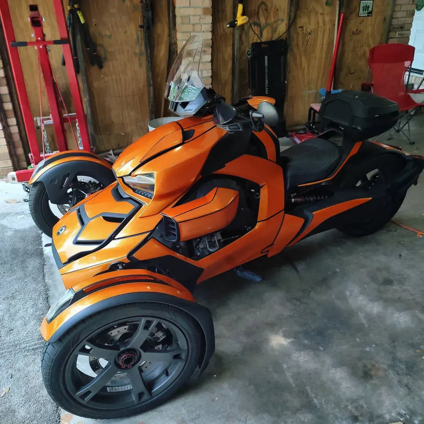 Orange and black Can-Am Spyder motorcycle parked inside a garage. Three-wheeled vehicle with a black storage box.