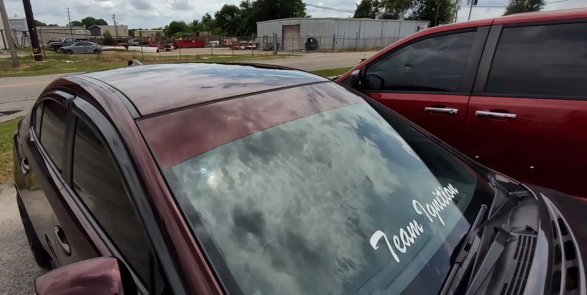 A maroon car with a windshield sticker parked next to a red car. Cloudy sky in the reflection.