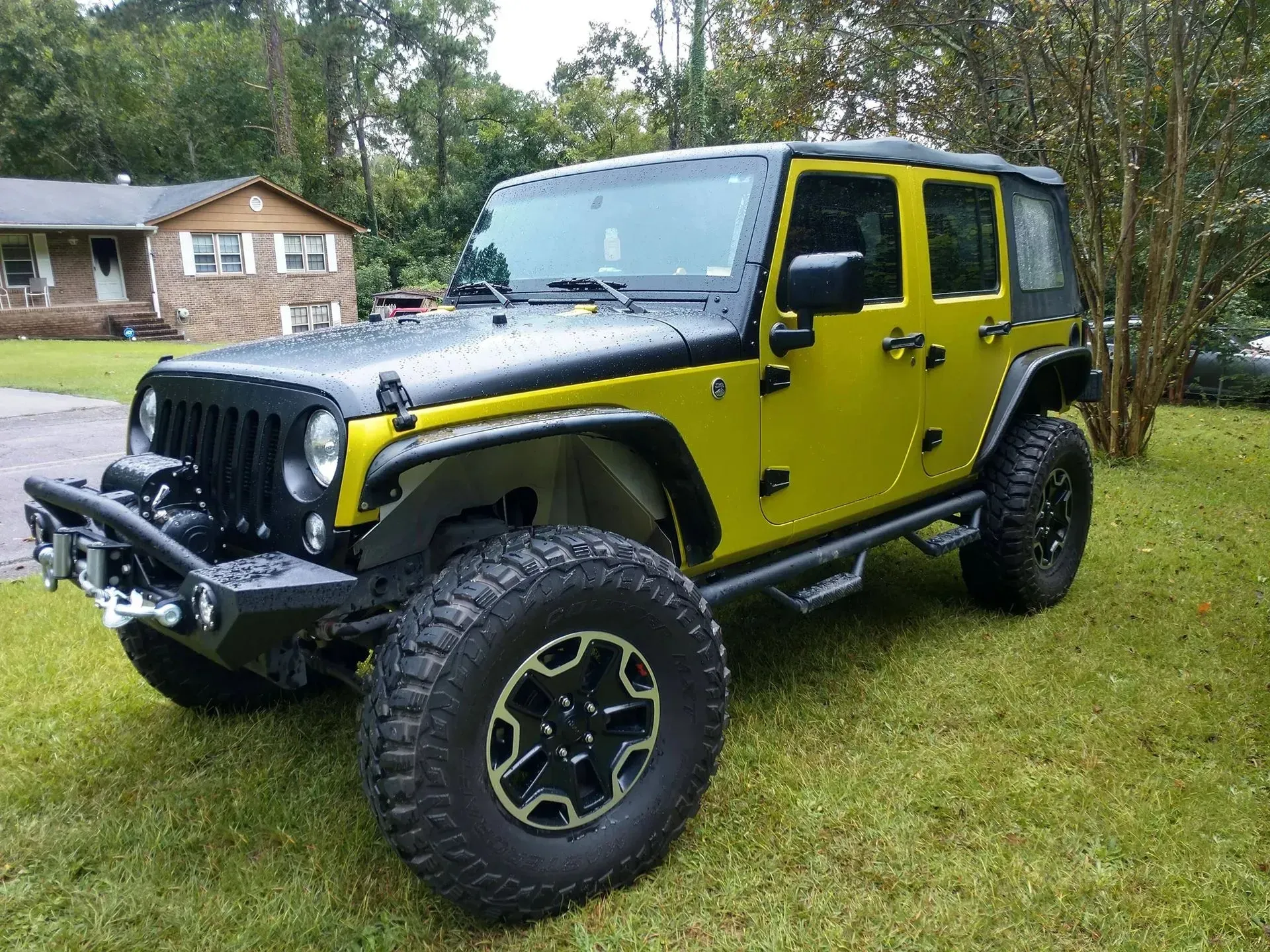 Yellow and black Jeep Wrangler with large off-road tires, parked on grass in front of a house.
