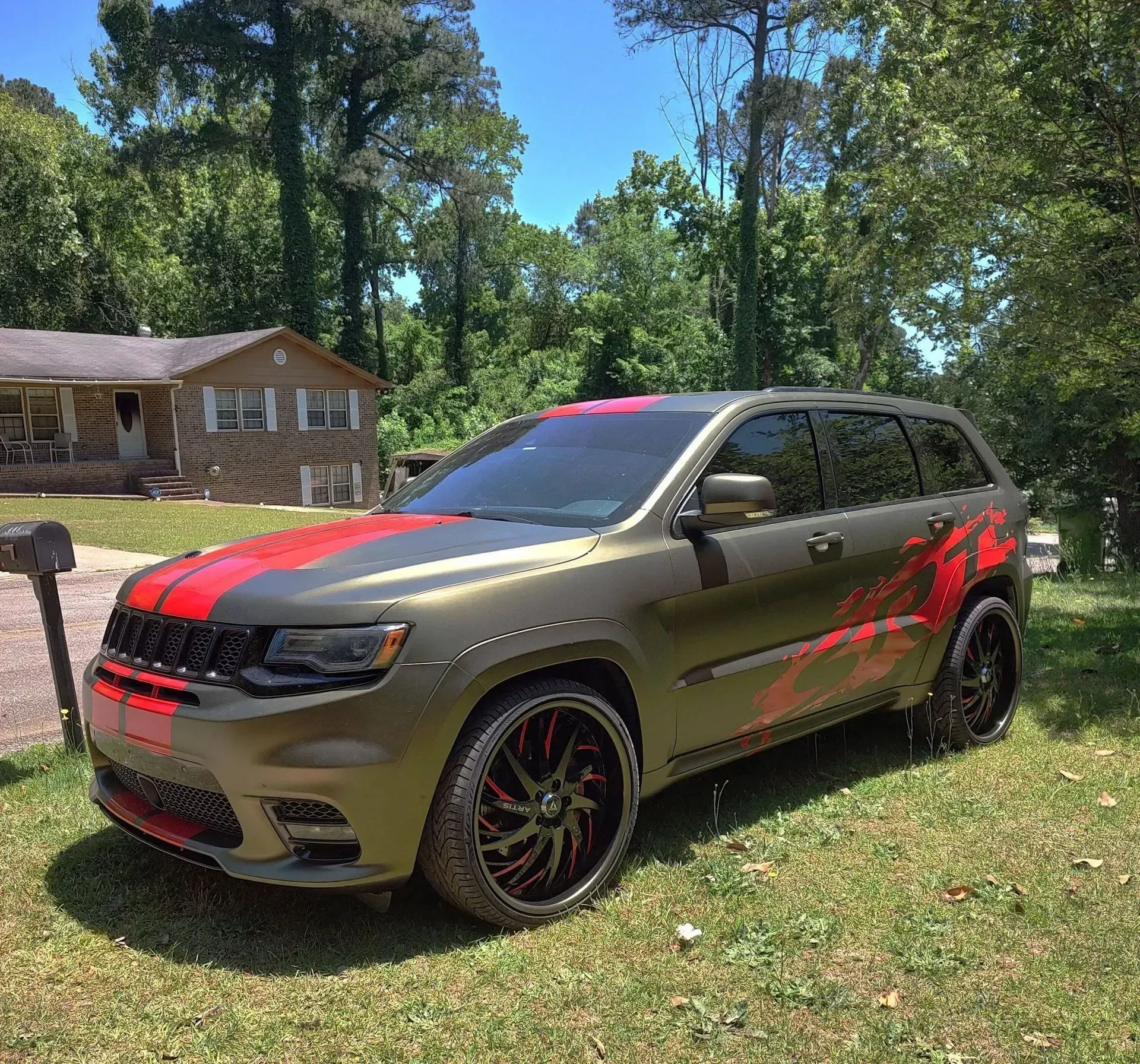Olive green SUV with red accents, parked in front of a house on a sunny day.