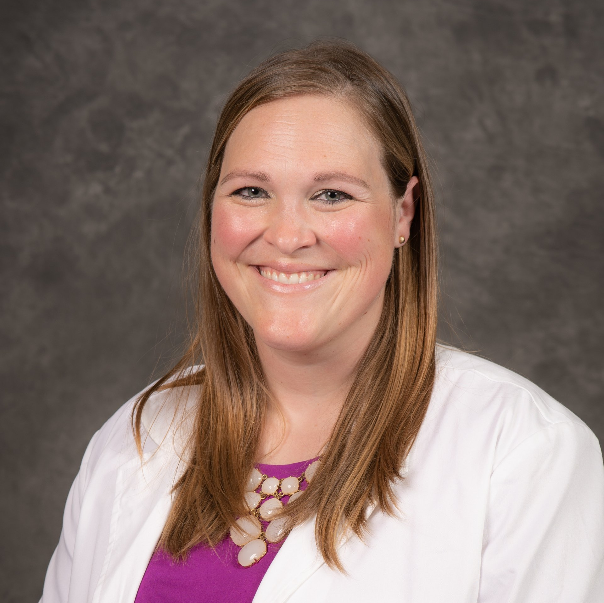 A woman in a white coat and purple shirt is smiling for the camera.