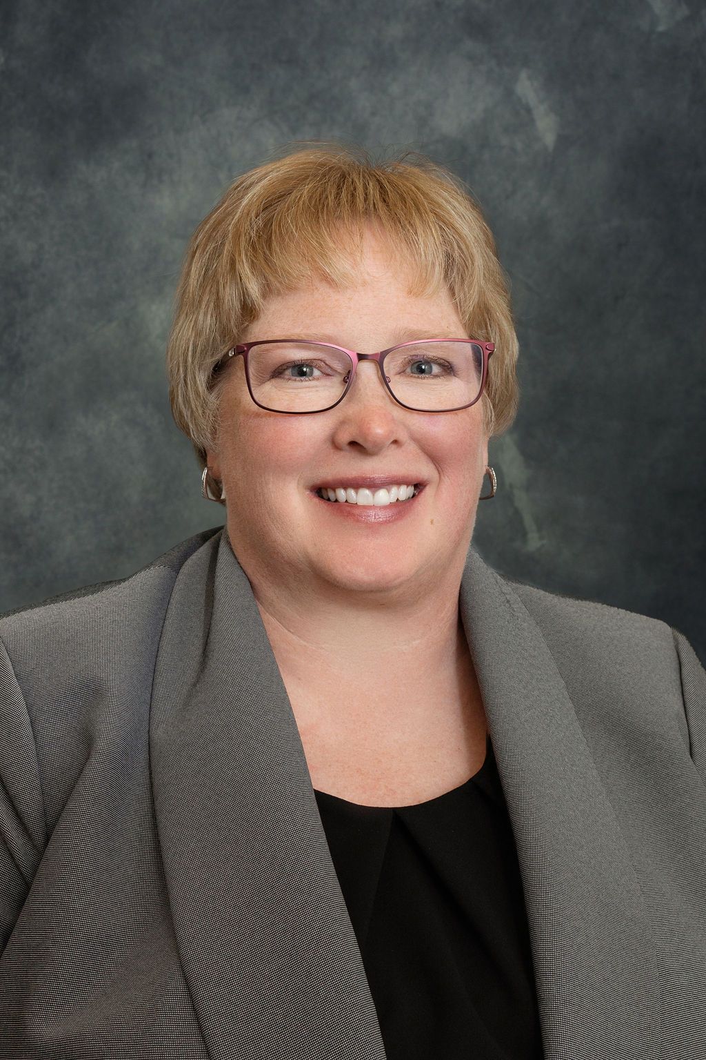 A woman wearing glasses and a gray jacket is smiling for the camera.