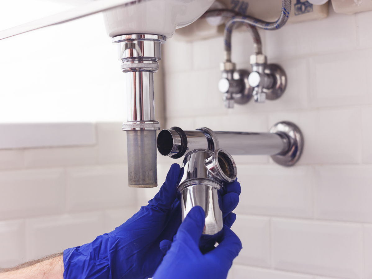 Plumber in blue gloves installing a chrome sink trap under a white sink.