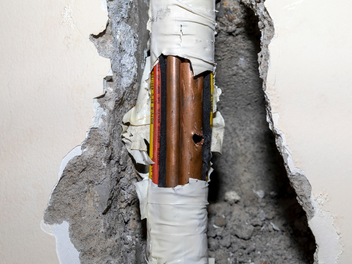 Damaged copper pipe in a concrete wall, with white tape and visible hole.