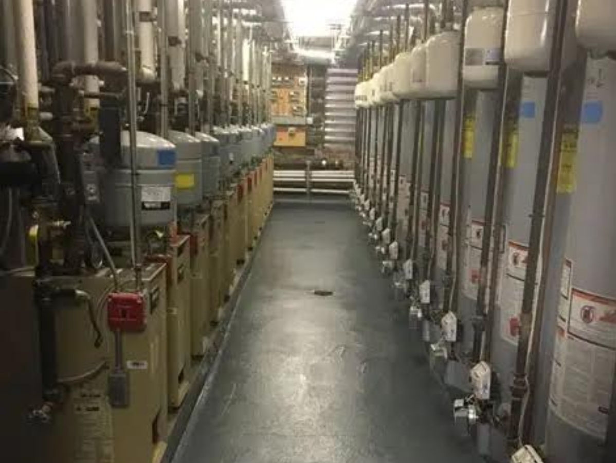 Narrow utility room with rows of water heaters and pipes.