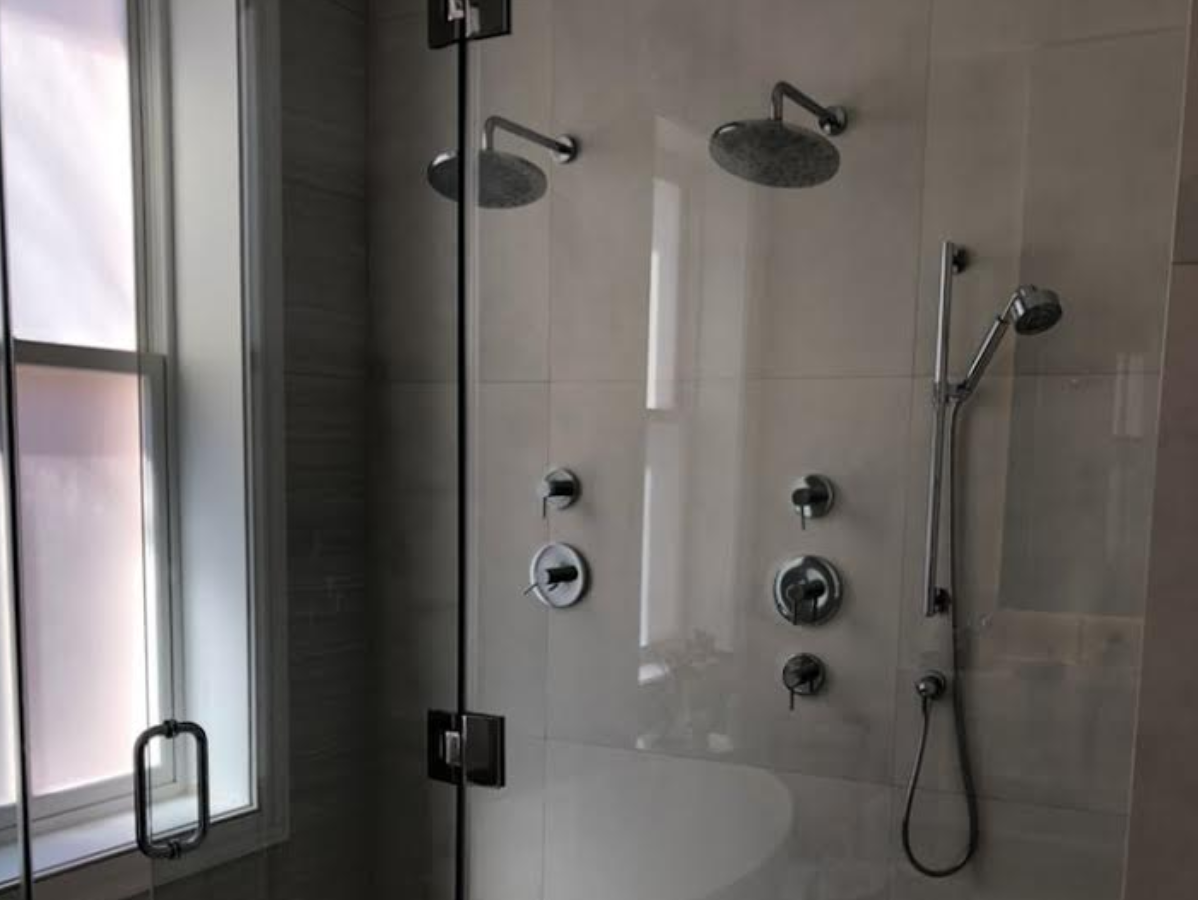 Glass-enclosed shower with two shower heads and a handheld shower, next to a window.
