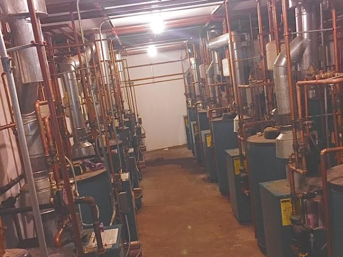 Rows of industrial boilers in a mechanical room, connected by pipes and wiring.