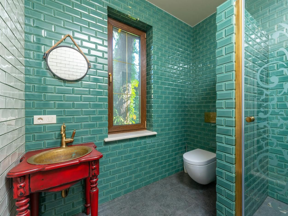 Teal tiled bathroom with red vanity, bronze sink, and window overlooking greenery.