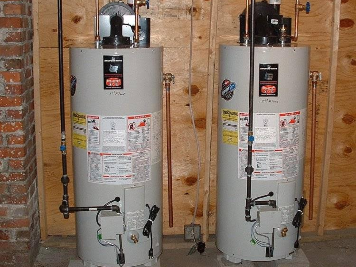 Two cylindrical water heaters stand side-by-side against a wooden wall. Copper pipes and various gauges are visible.