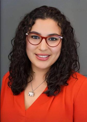 Portrait of a smiling woman with curly hair, red glasses, and a bright red top against a gray background