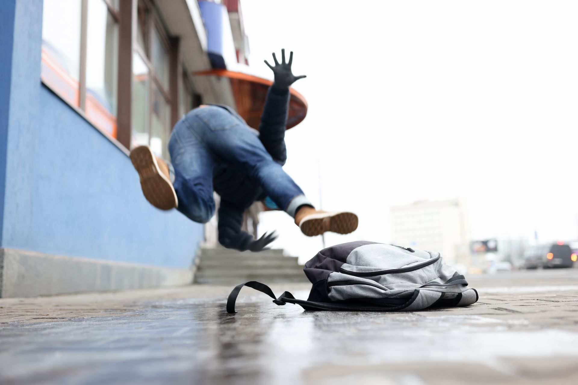 Person falling on slick pavement next to backpack. Hands raised, blue building in background.
