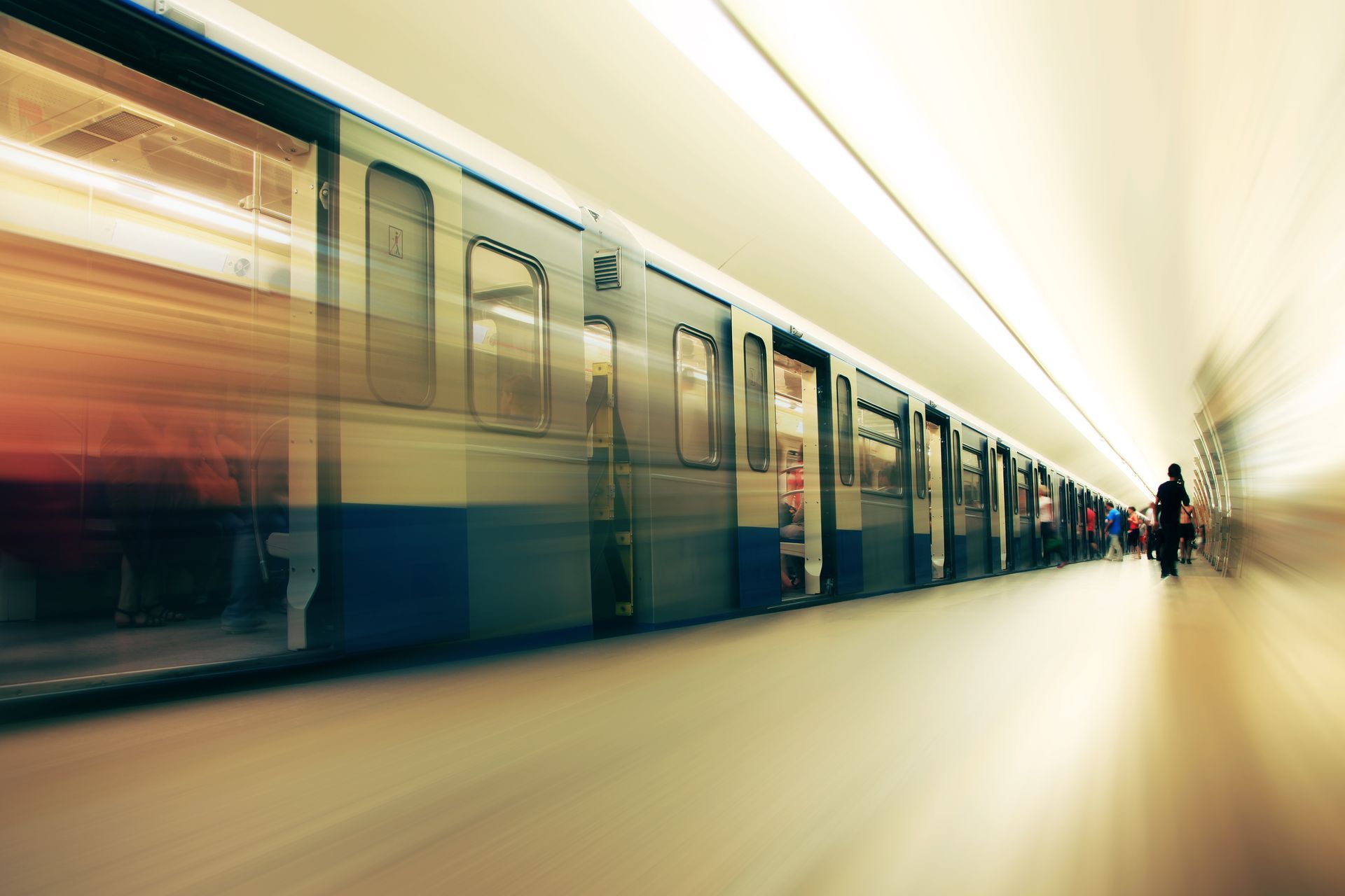 Subway train speeding in a station; blurred motion, passengers, white and blue train cars, linear perspective.