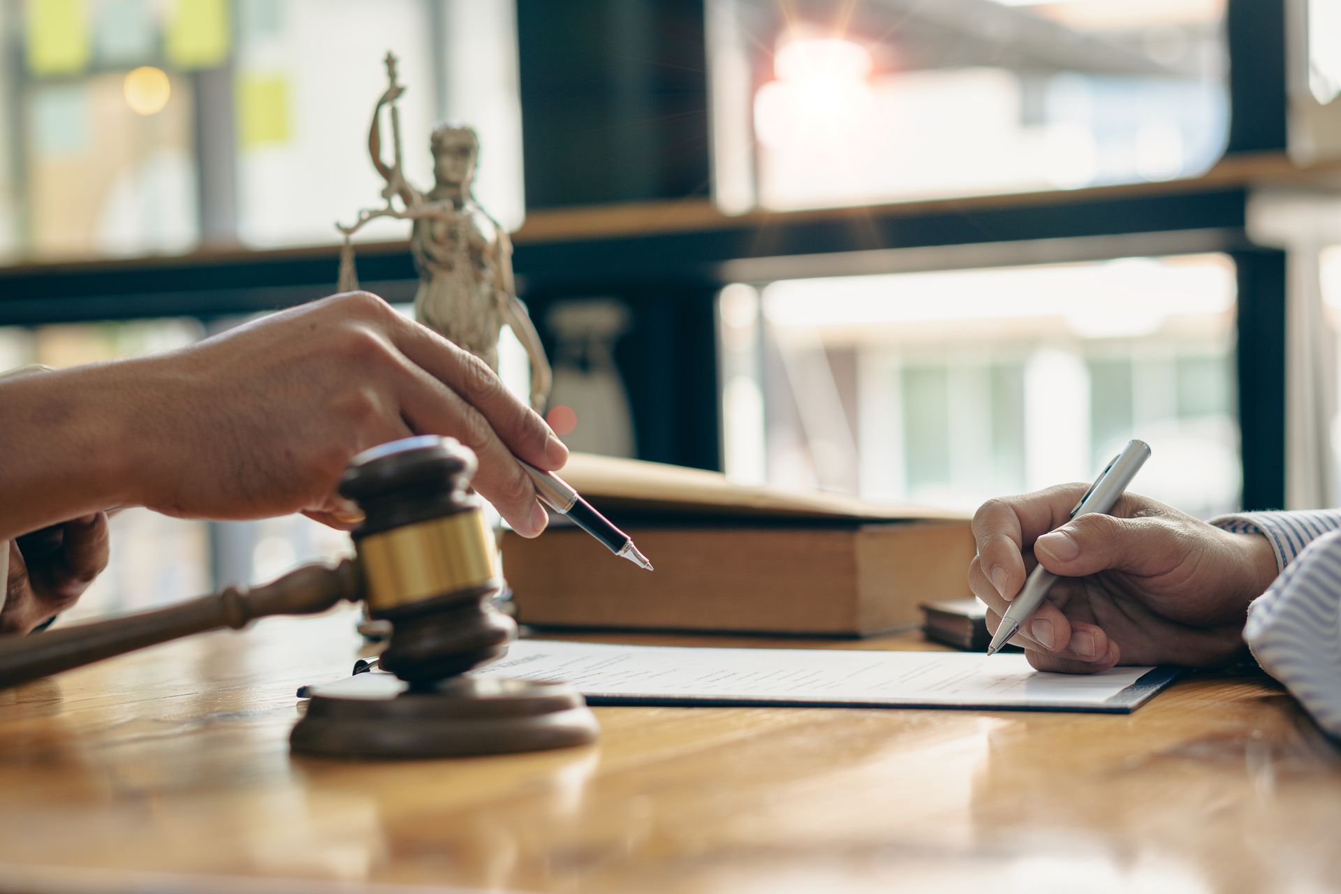 Two people sign a document at a table with a gavel and Statue of Justice in the background.