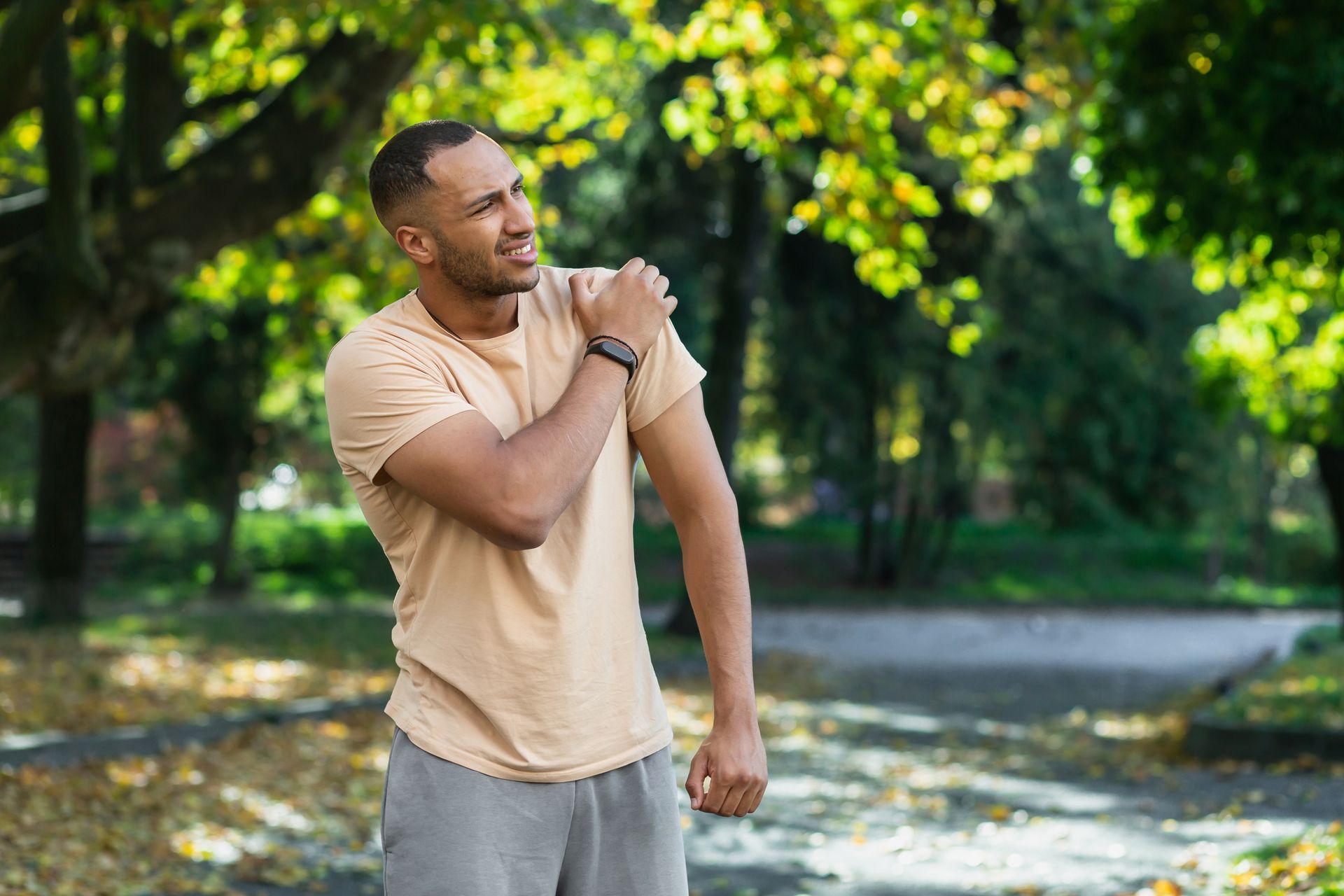 Man in beige shirt touching his shoulder in pain, outdoors in a park.