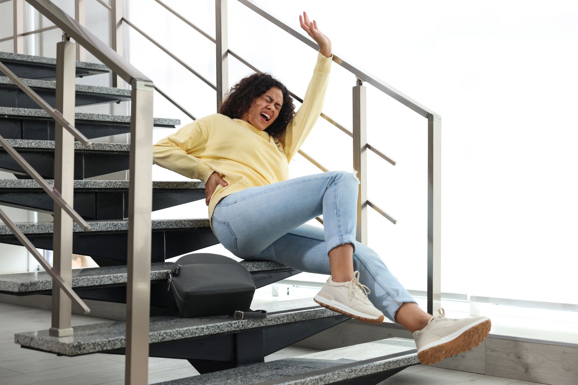 Woman falling down a staircase, clutching her back. She wears jeans, a yellow shirt, and sneakers.