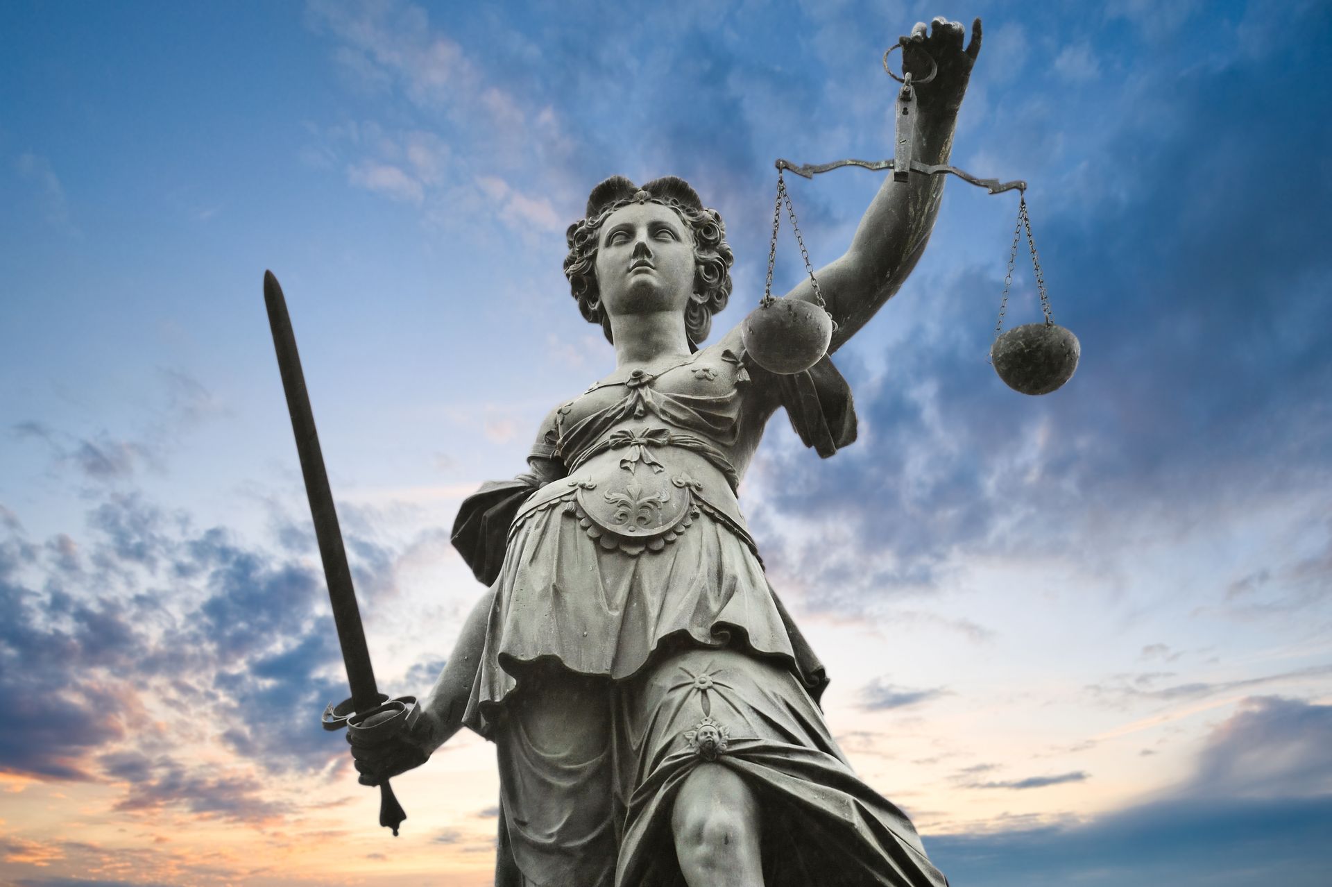 Statue of Lady Justice holding scales and sword, set against a cloudy sky.