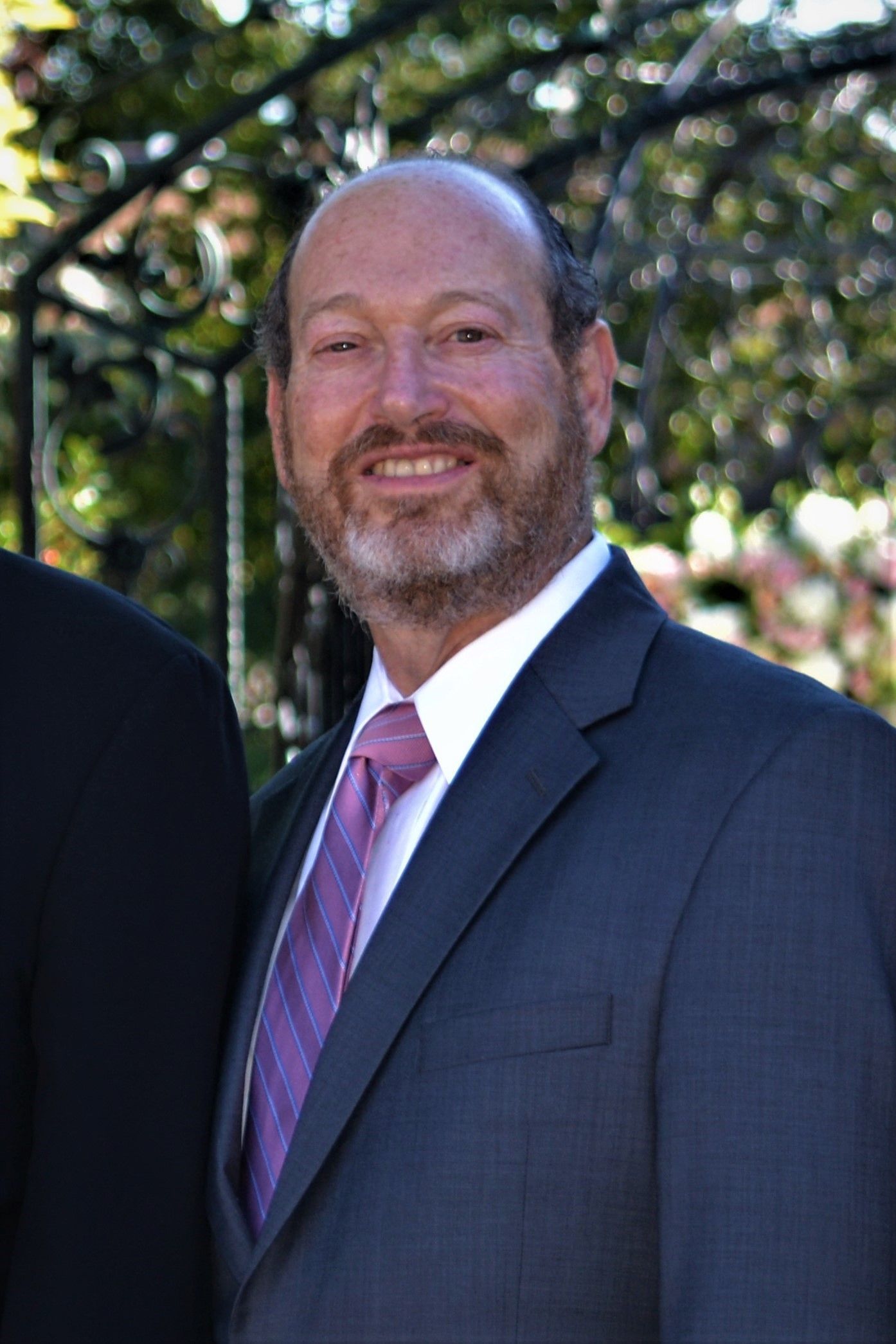 Man with a beard wearing a suit and tie, smiling, outdoors with greenery.