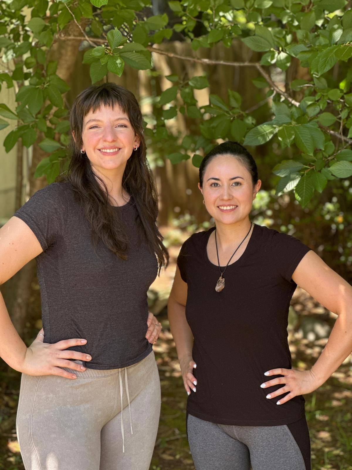 Two women, hands on hips, smiling in front of green foliage. One wears grey, the other black.