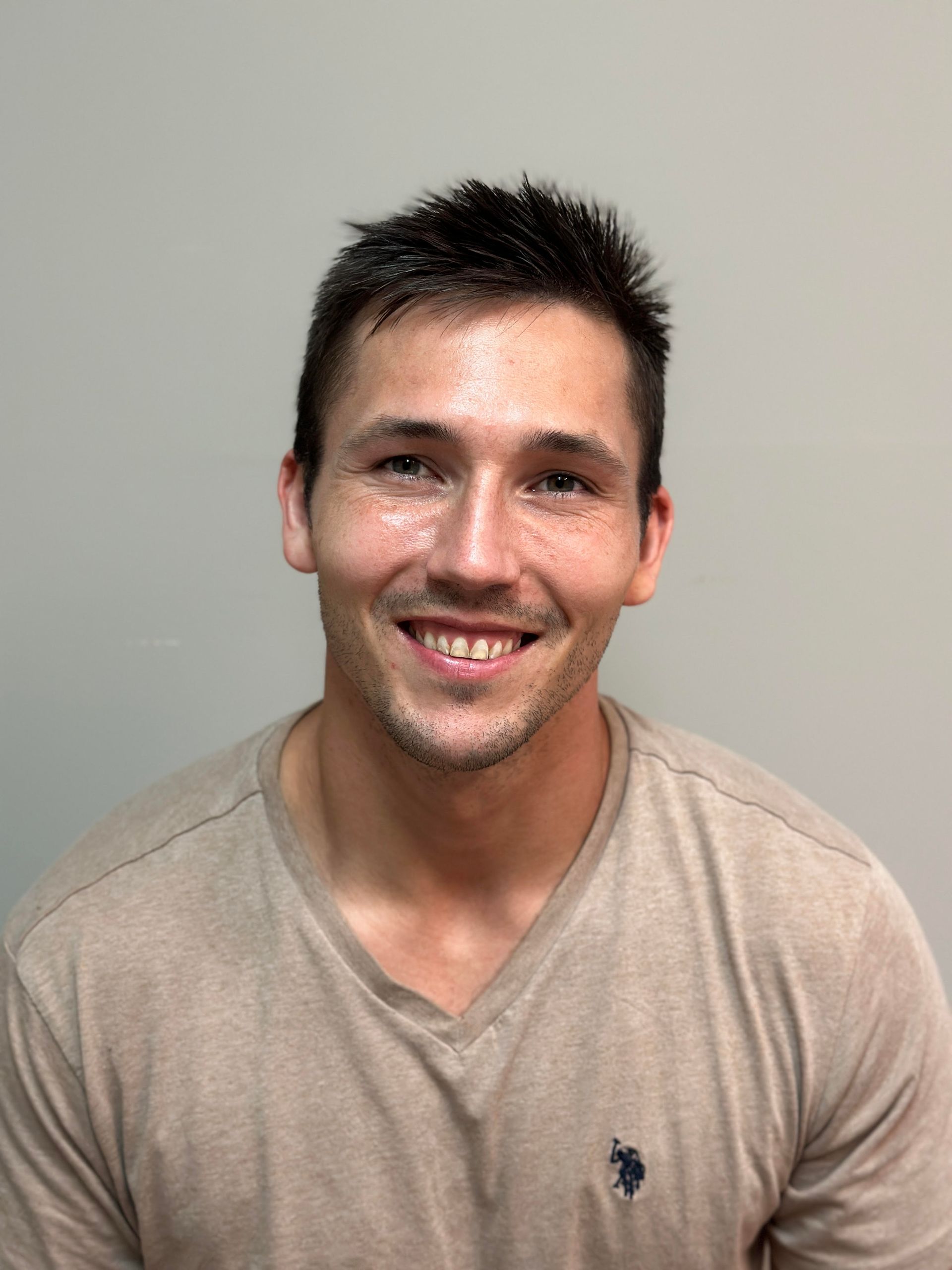 Man smiling, wearing a beige v-neck shirt, in front of a neutral wall.