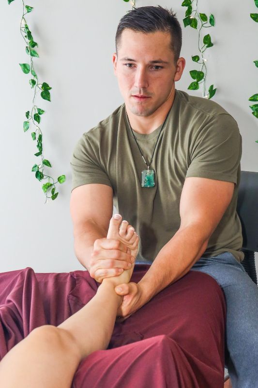Man giving a foot massage, holding foot on a cloth-covered surface. Light-colored walls with vines in the background.
