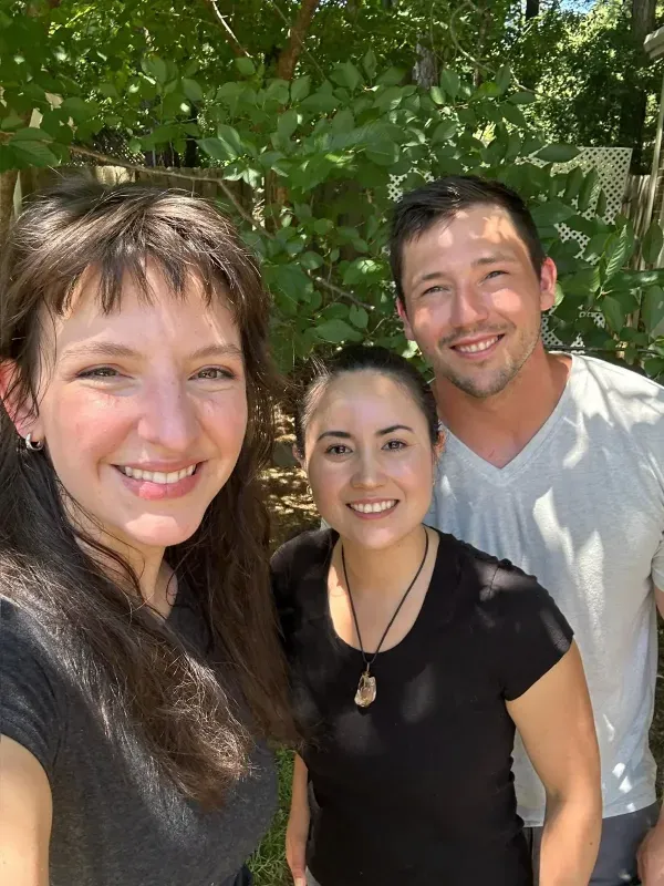 Three smiling people pose outside in front of green foliage.