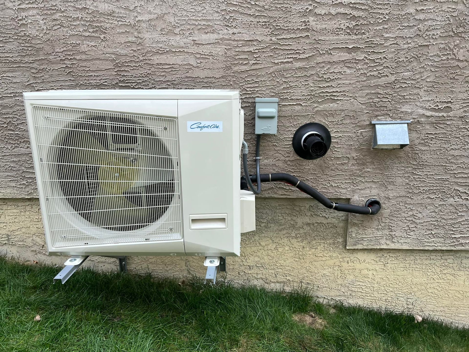 Outdoor heat pump mounted on a stucco wall above grass. Beige unit, black piping, electrical boxes.