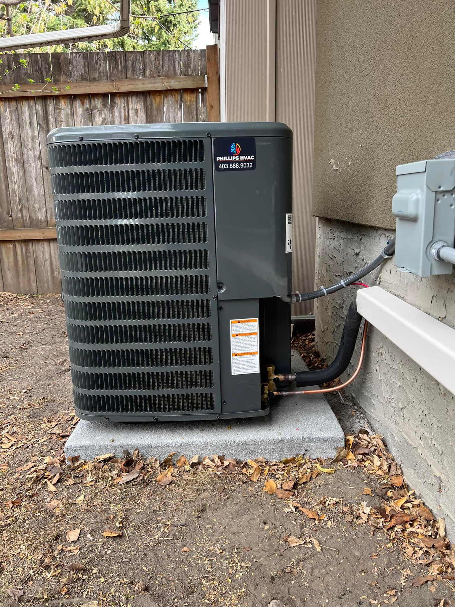 An air conditioning unit on a concrete base, near a house and a wooden fence.