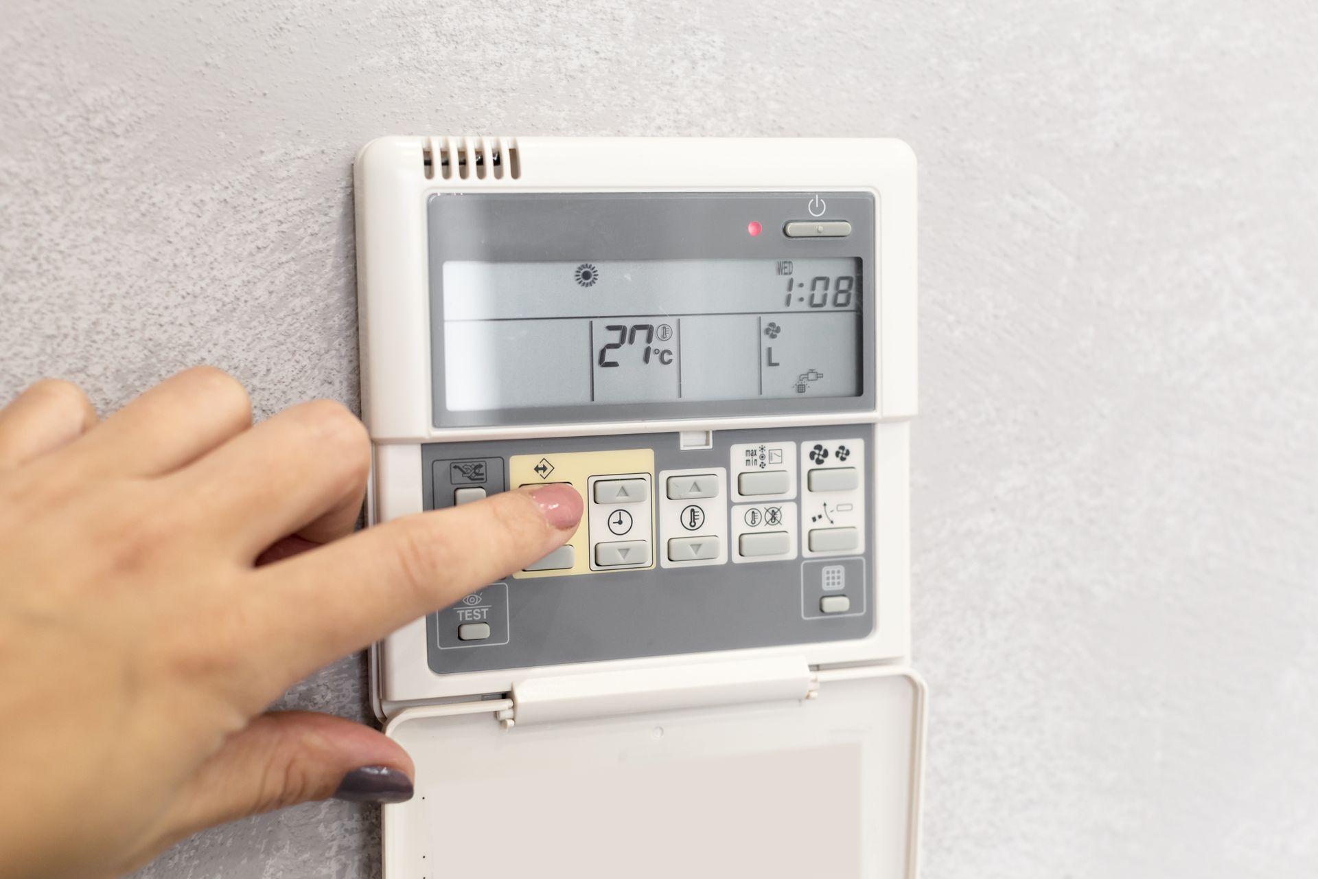 Hand pressing a button on a white thermostat mounted on a gray wall. The display shows the temperature.