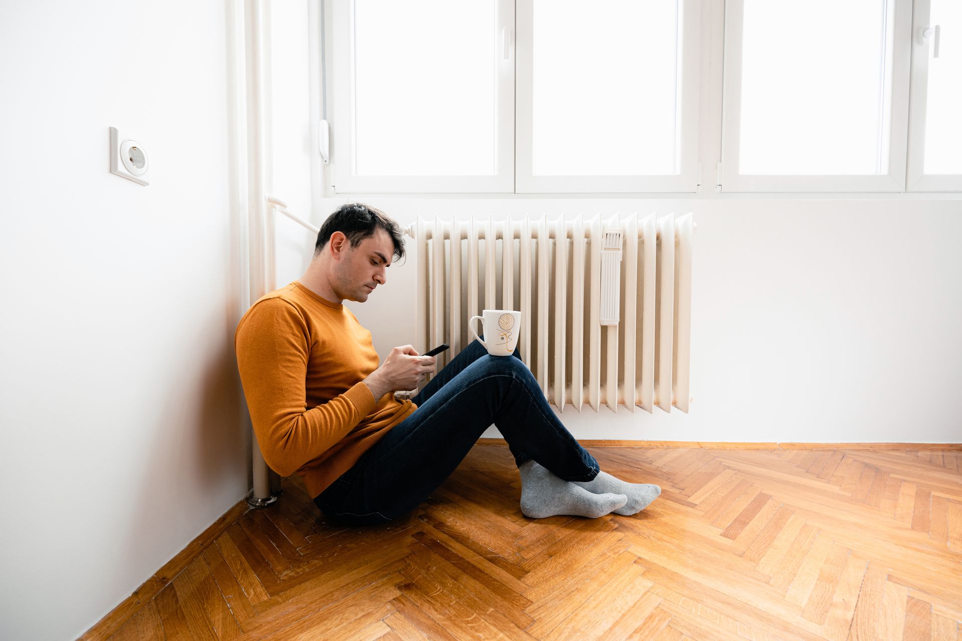 A man is sitting on the floor with a cup of coffee and looking at his phone.