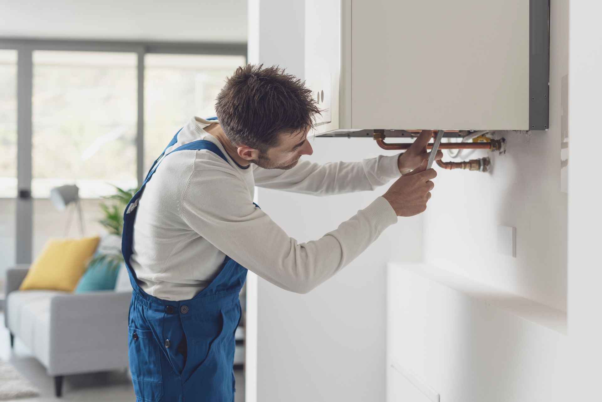 A man is fixing a boiler on a wall in a living room.