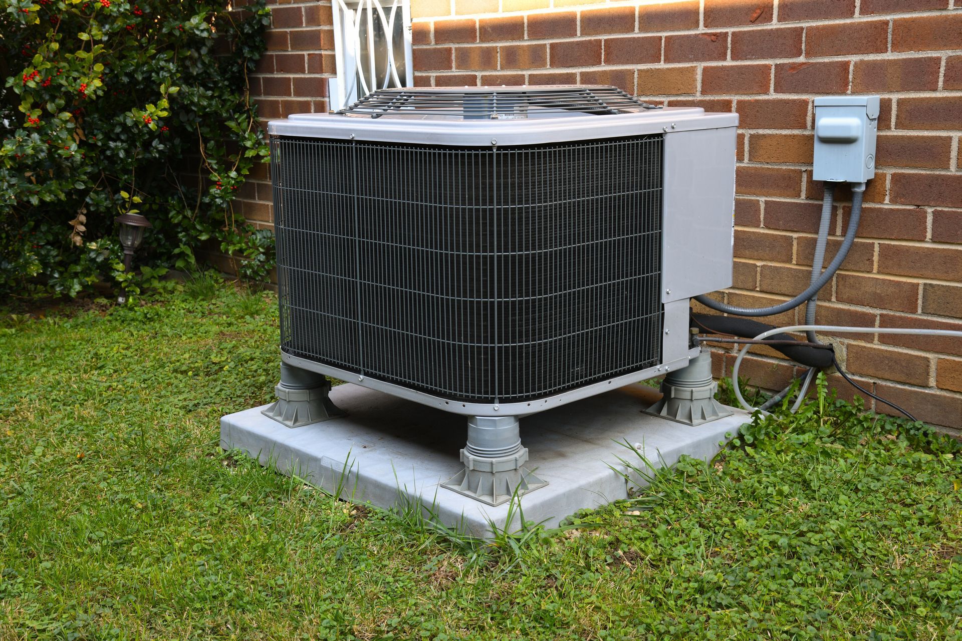 An air conditioner is sitting in the grass next to a brick wall.