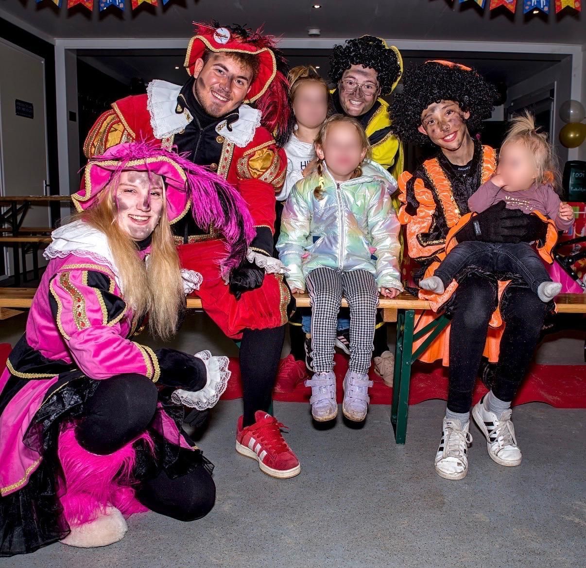 Een groep mensen in kleurrijke Halloweenkostuums poseert binnen, met twee kleine kinderen vooraan.