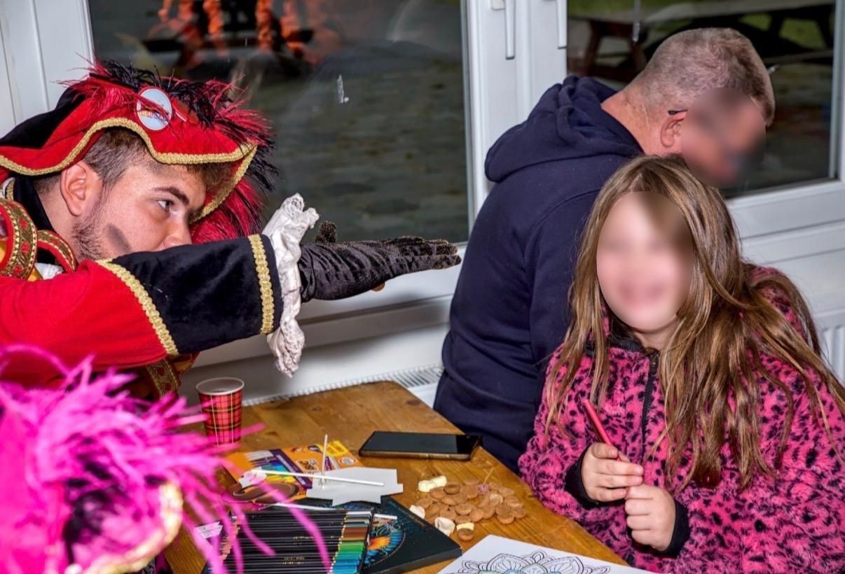 Een artiest in kostuum wijst naar een kind dat aan een tafel zit met knutselspullen en papier in een lichte binnenruimte.