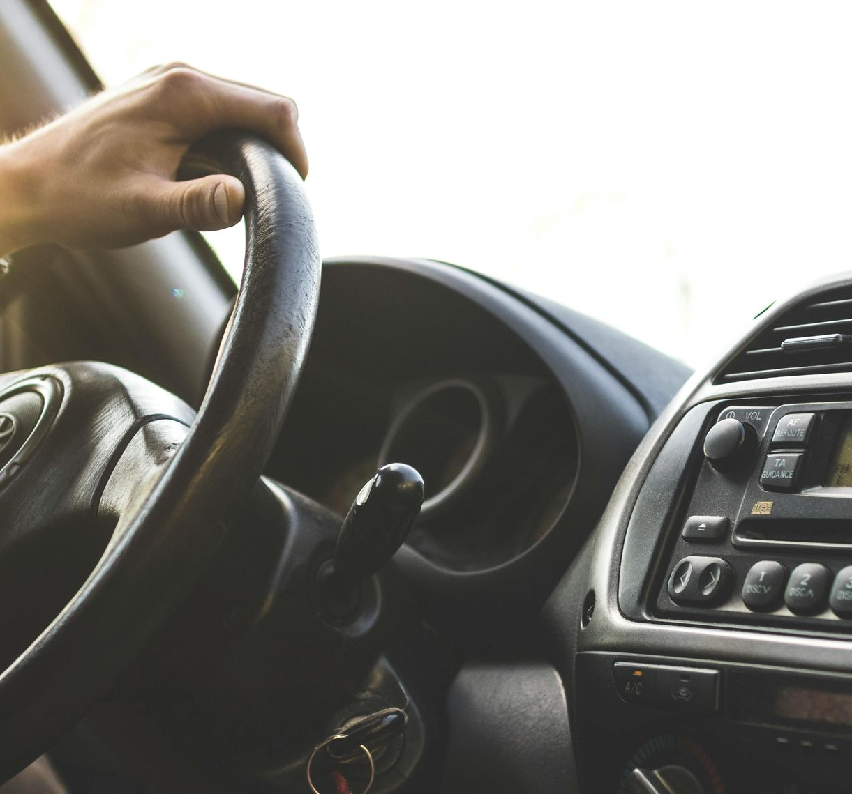 Hand gripping a car steering wheel, dashboard visible. Sunlight.