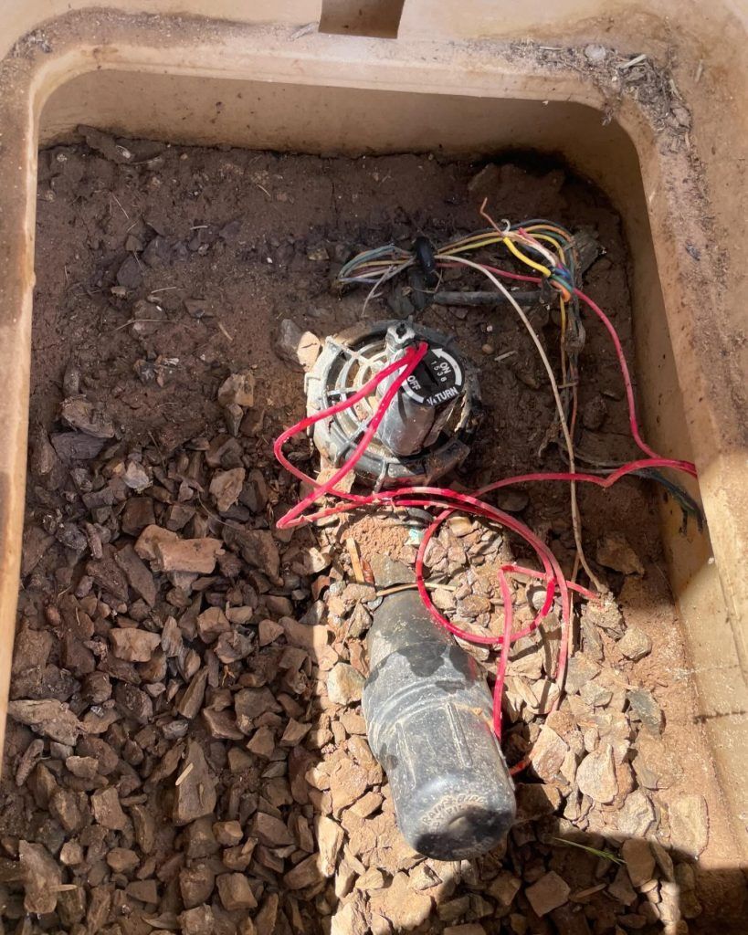 An open irrigation valve box with sprinkler system components and exposed wires. Soil and rocks fill the box.