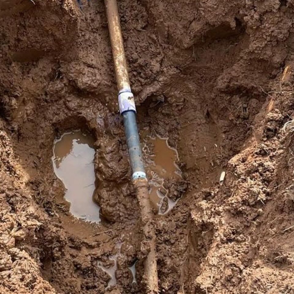 A broken pipe in a muddy hole with standing water, revealing a repair coupling.